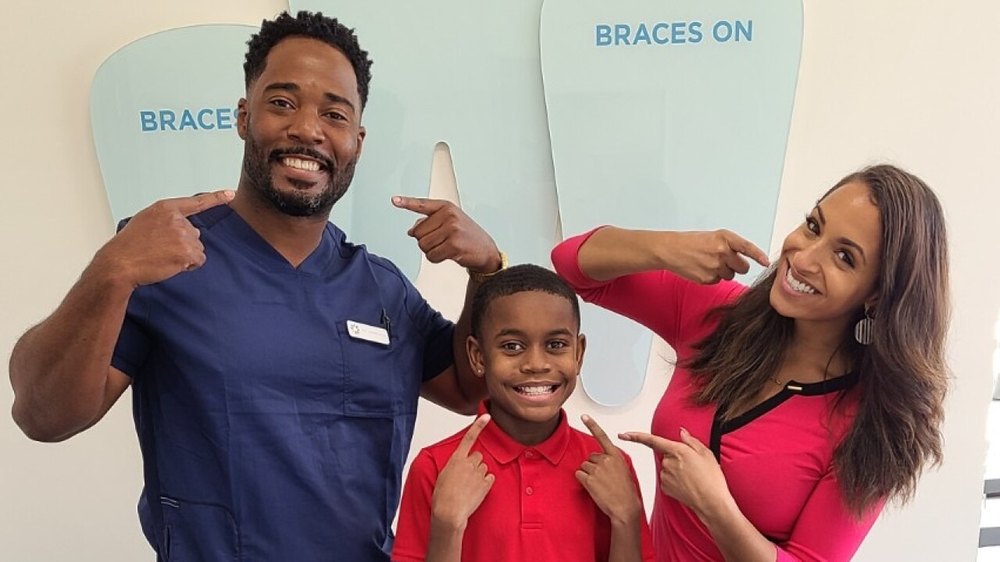 Dr. Ray Tucker and a colleague pointing at their smiles beside a young patient in front of a Braces On display